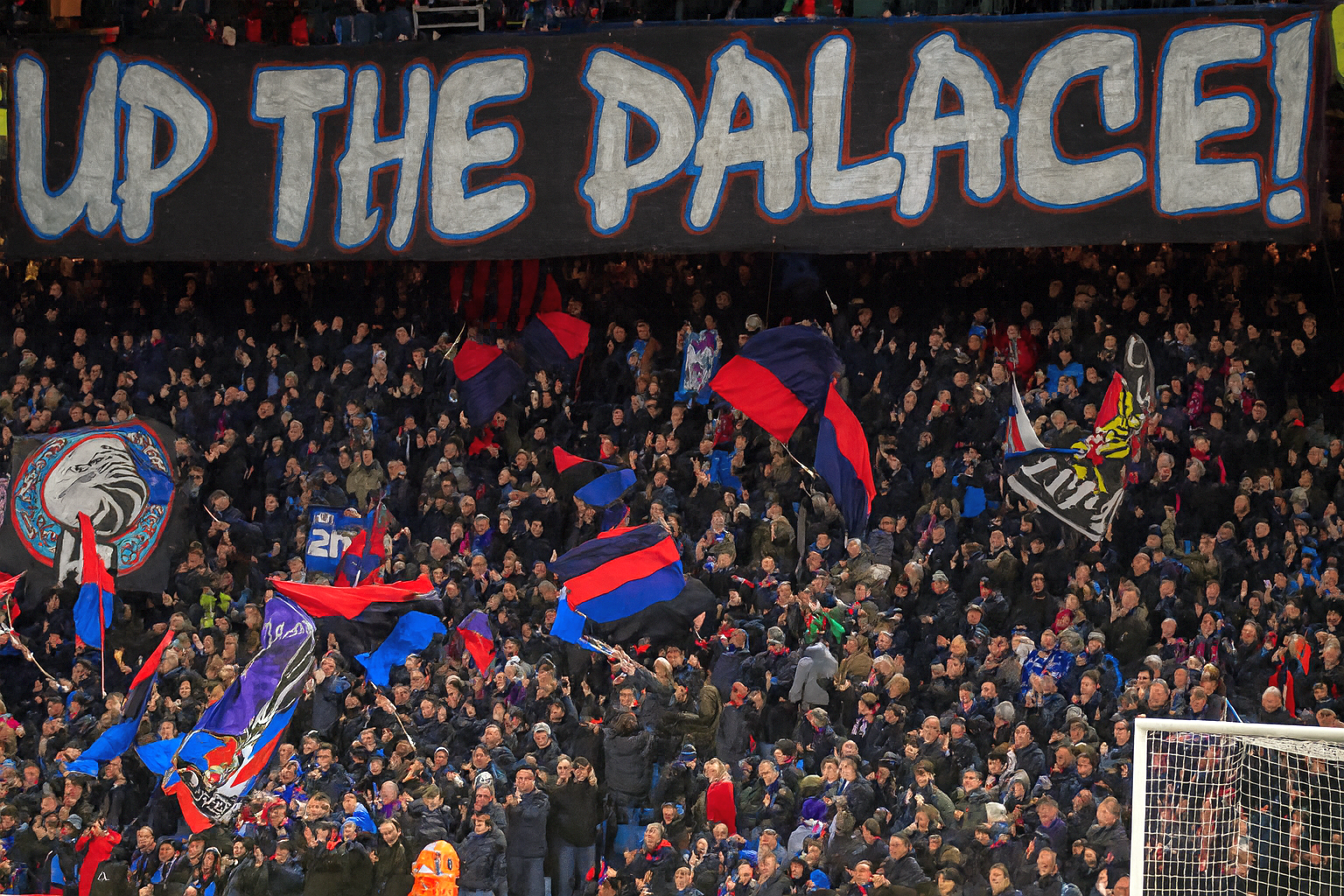 En stor folkmassa av fotbollsfans på en stadion med en banderoll som lyder "UP THE PALACE!" ovanför dem, många viftar med lagflaggor.
