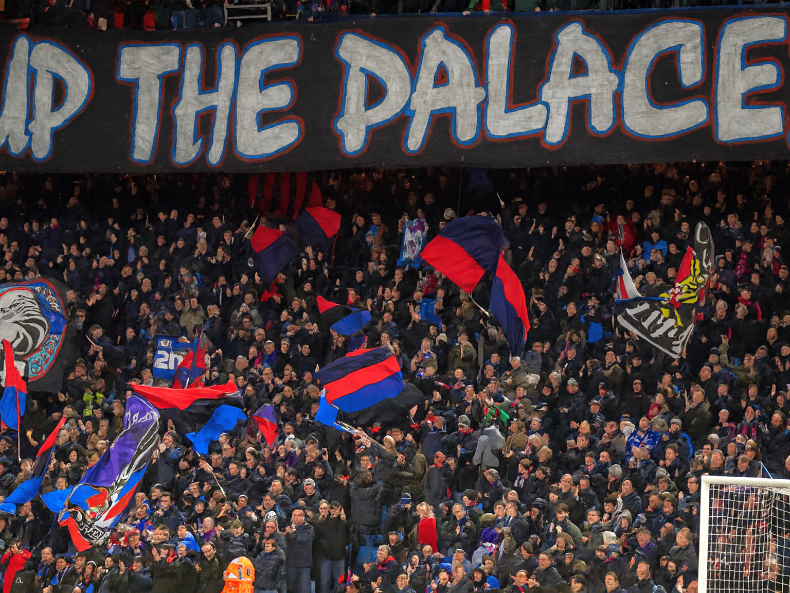 A large crowd of football fans in a stadium with a banner reading "UP THE PALACE!" above them, many waving team flags.