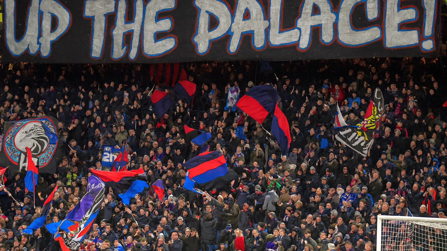 En stor folkmassa av fotbollsfans på en stadion med en banderoll som lyder "UP THE PALACE!" ovanför dem, många viftar med lagflaggor.
