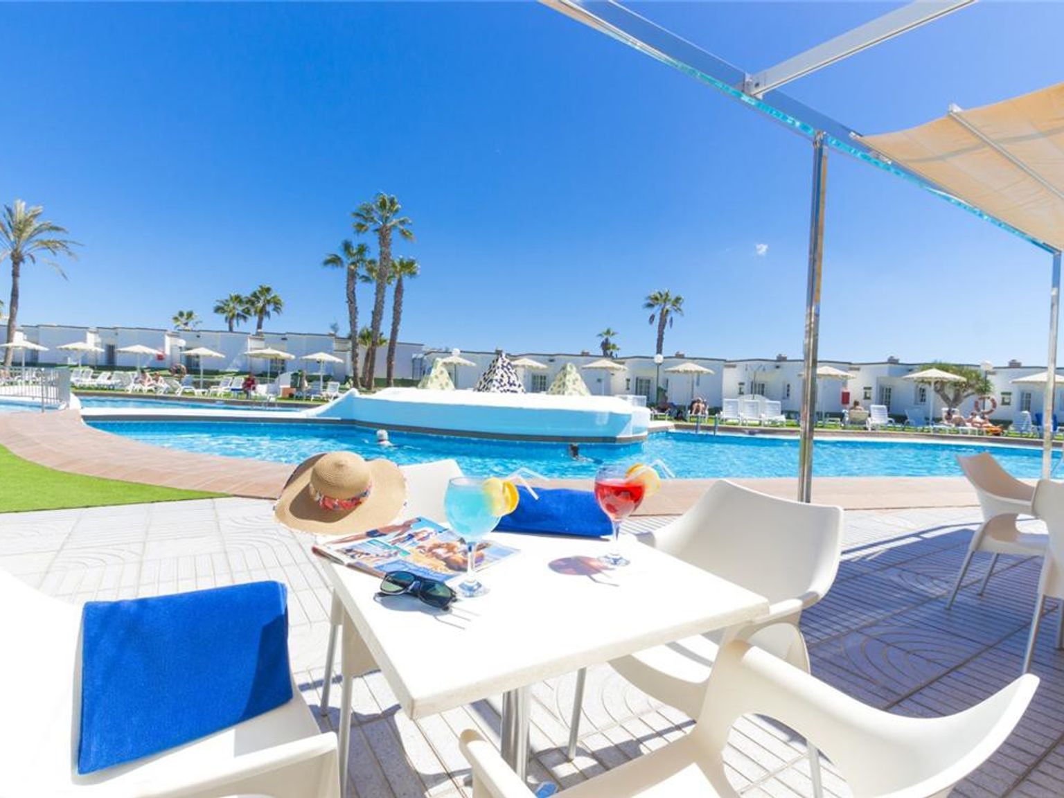 En resortterrasse med cocktails, en hatt og magasiner på et bord, med utsikt over et stort svømmebasseng, hvite bungalower og palmer under en klar blå himmel.