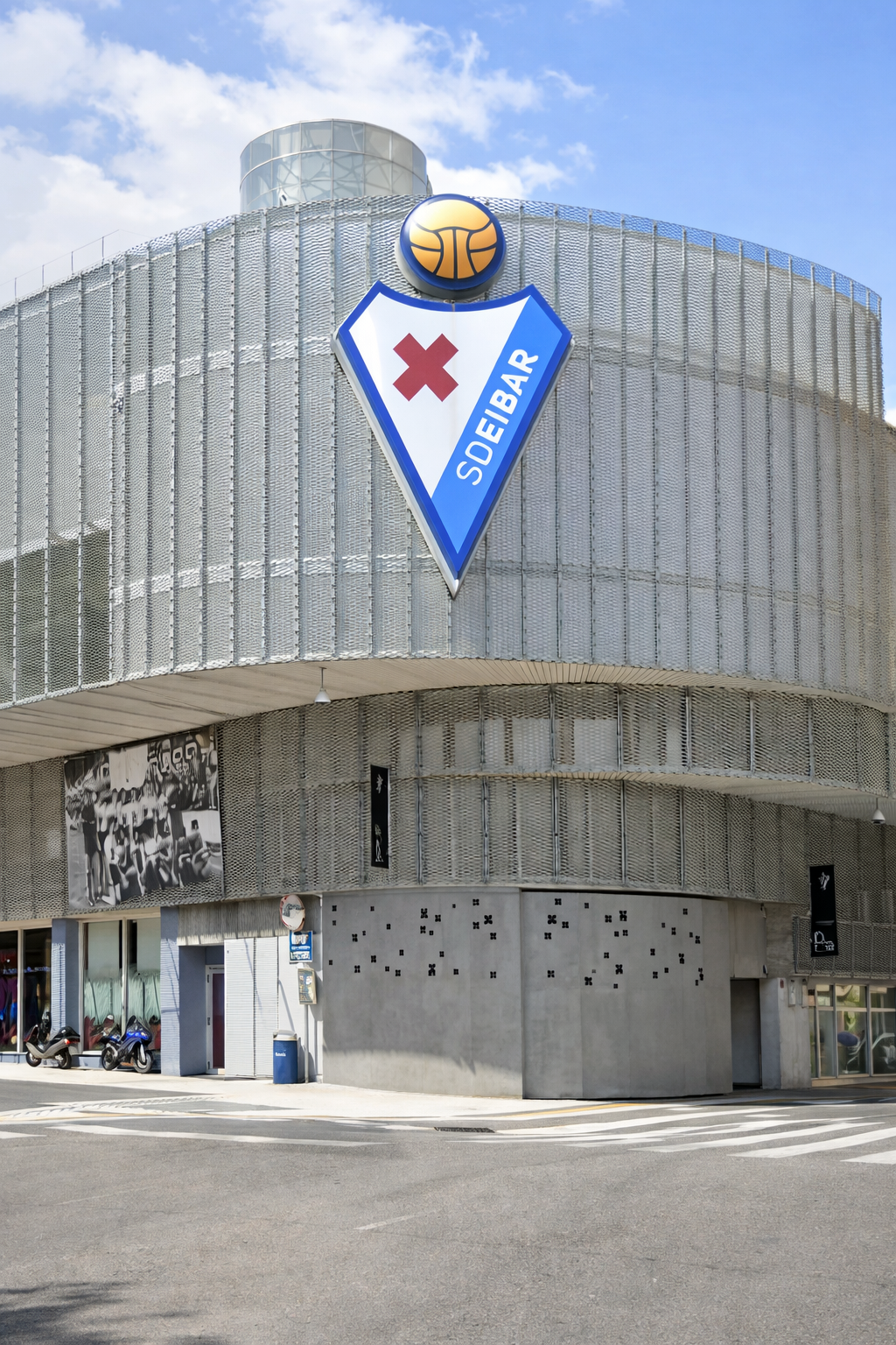 Modern building with a metal mesh facade, featuring a large triangular "SDEIBAR" logo with a red cross and a basketball icon on top, next to a street.