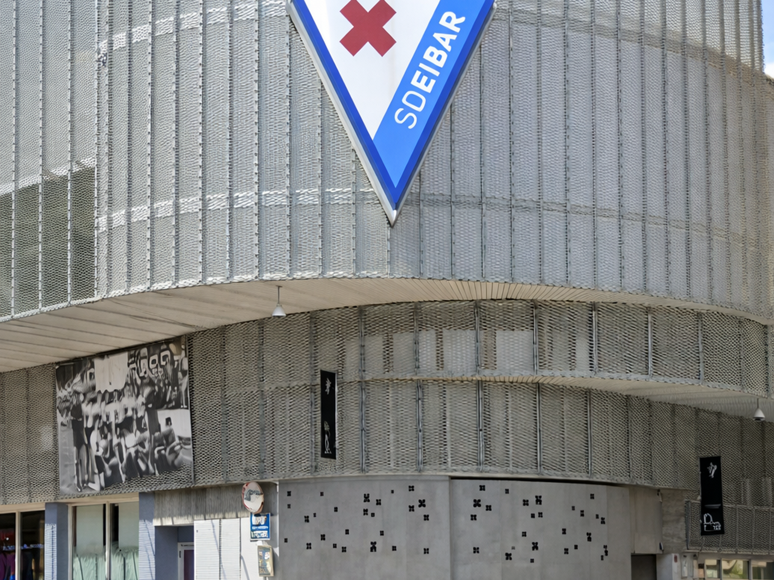 Modern building with a metal mesh facade, featuring a large triangular "SDEIBAR" logo with a red cross and a basketball icon on top, next to a street.