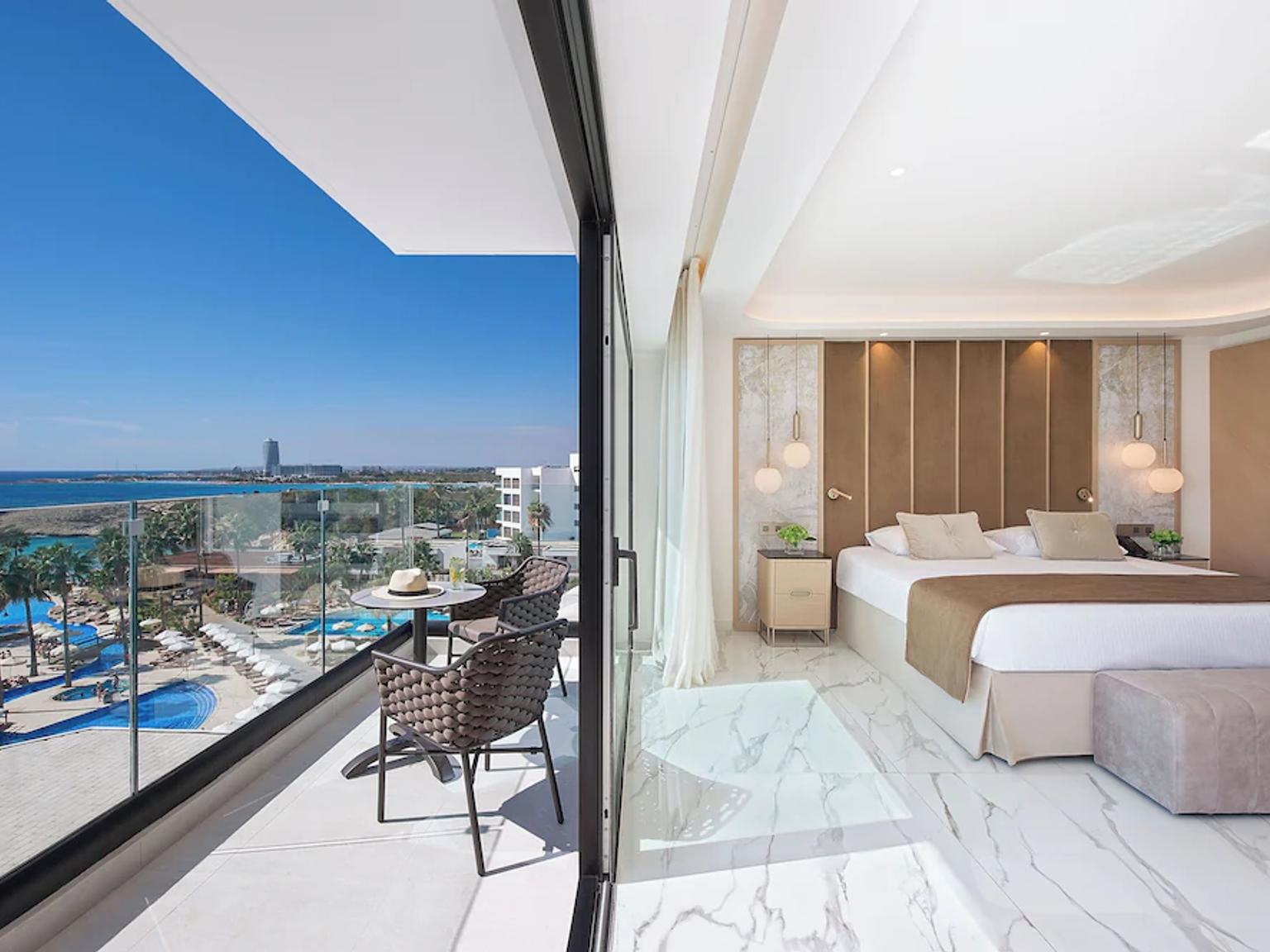 Et moderne hotellrom med en stor seng og en balkong med utsikt over et resortbassengområde og havet.