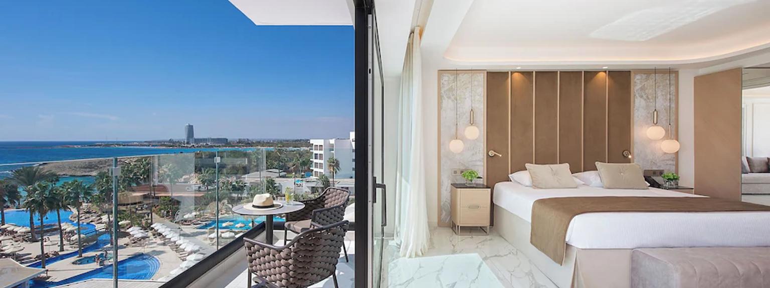 Et moderne hotellrom med en stor seng og en balkong med utsikt over et resortbassengområde og havet.