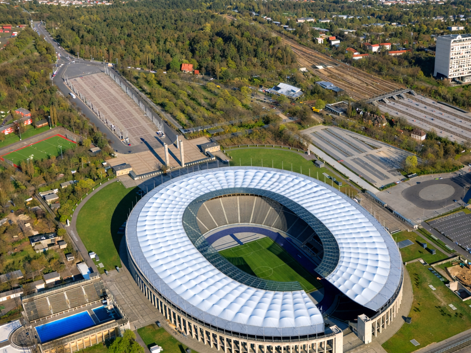 Luftaufnahme des Olympiastadions Berlin, ein ovales Stadion mit einem weißen, geriffelten Dach, umgeben von grünen Parkanlagen und städtischen Gebieten.
