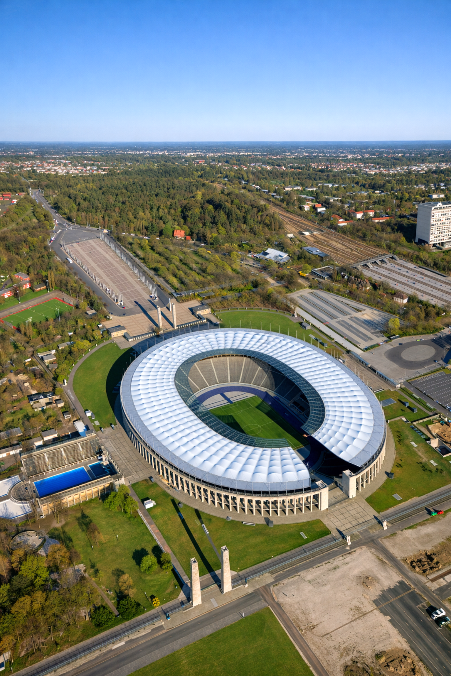 Luftaufnahme des Olympiastadions Berlin, ein ovales Stadion mit einem weißen, geriffelten Dach, umgeben von grünen Parkanlagen und städtischen Gebieten.