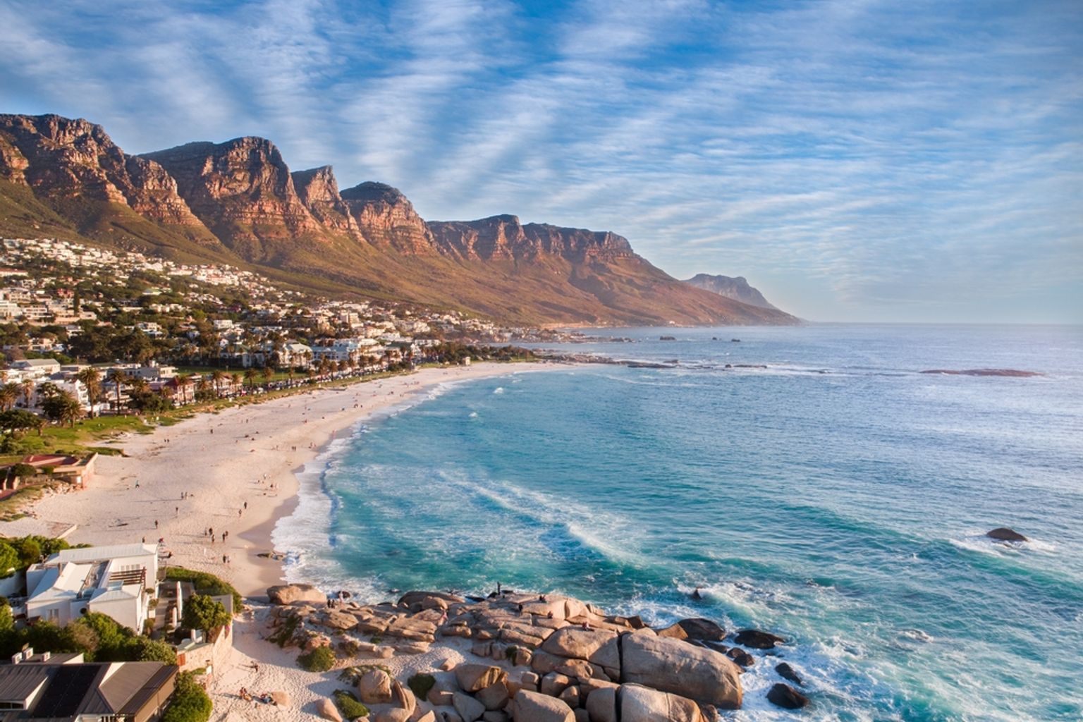 Luftfoto af Camps Bay i Cape Town med en bred sandstrand, turkis hav og bjerge.