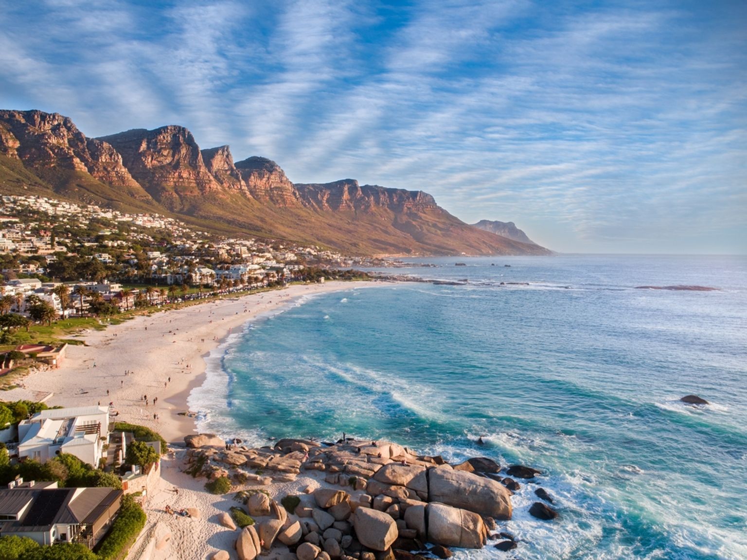Flygfoto över Camps Bay i Kapstaden, med en bred sandstrand, turkos ocean och berg.