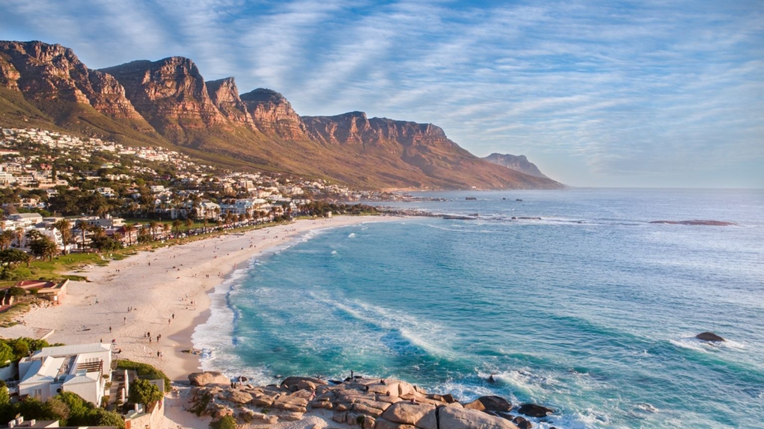 Luftfoto af Camps Bay i Cape Town med en bred sandstrand, turkis hav og bjerge.