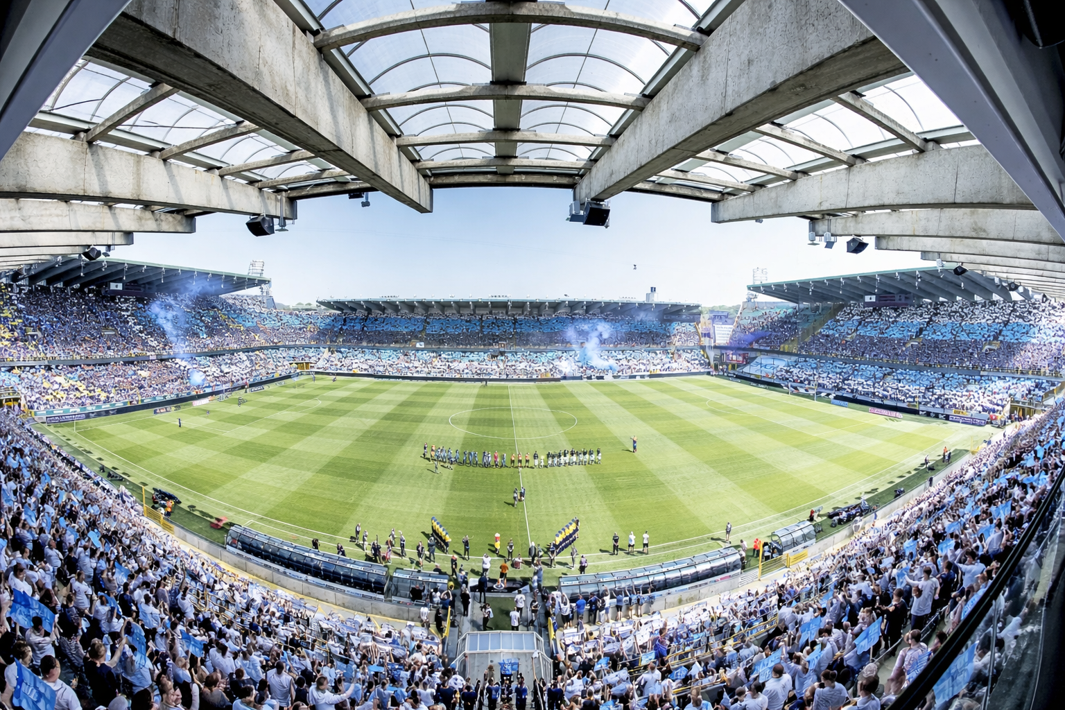 Wide shot of a packed football stadium with fans in blue and white, blue smoke, and players on the pitch.