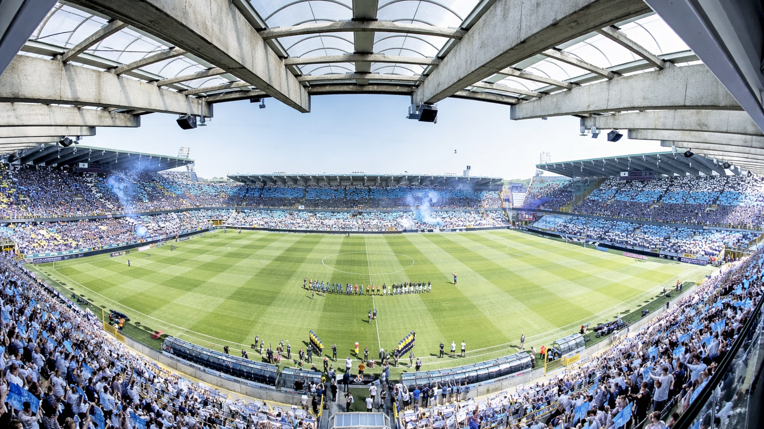 Wide shot of a packed football stadium with fans in blue and white, blue smoke, and players on the pitch.