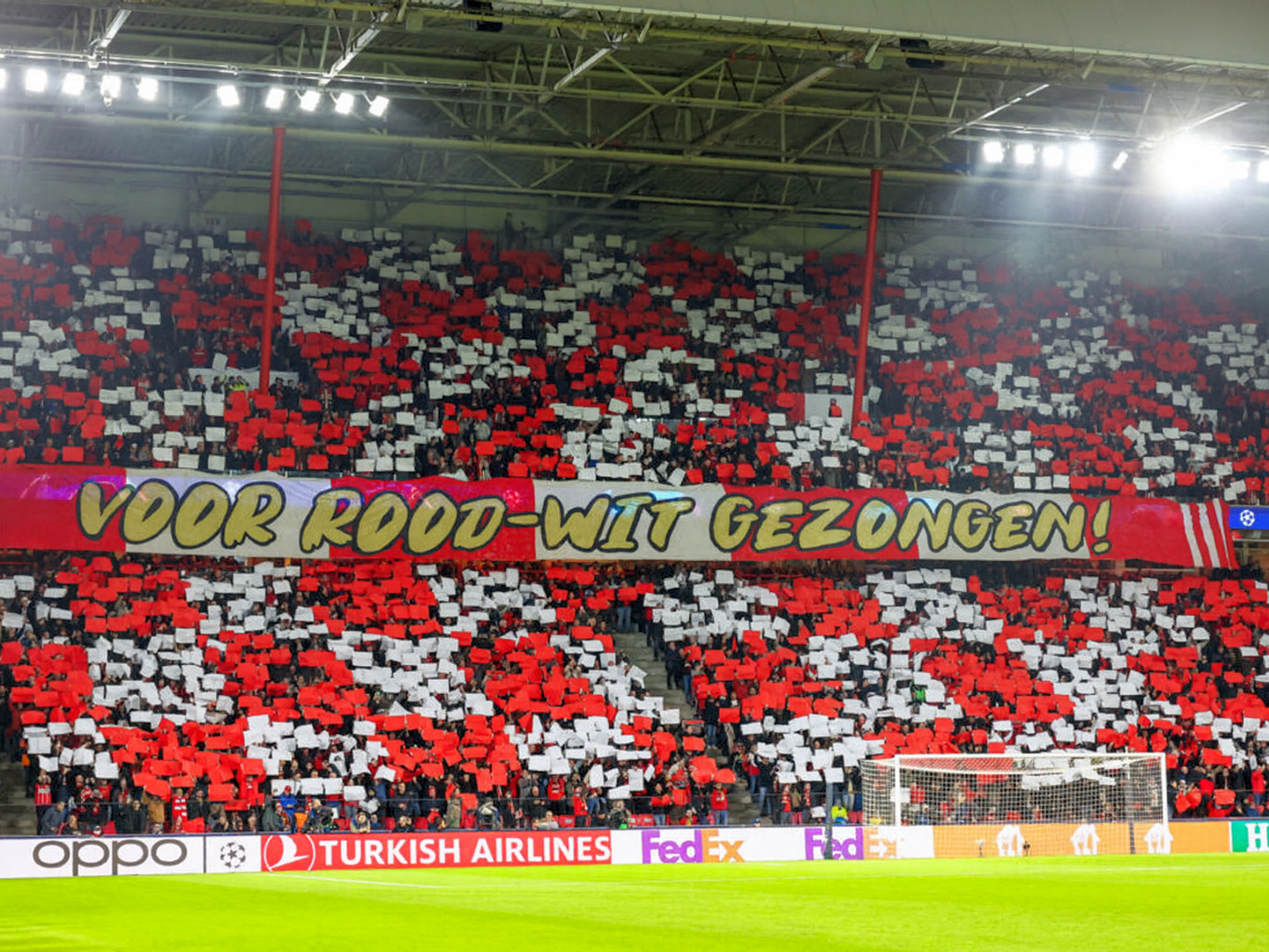 Fotbollsfans på en stadion håller upp röda och vita kort och skapar en display under en banderoll som lyder 'VOOR ROOD-WIT GEZONGEN!'.