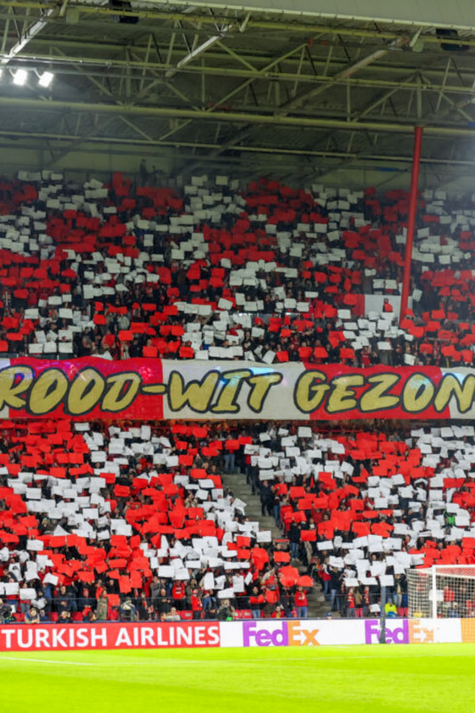 Fotbollsfans på en stadion håller upp röda och vita kort och skapar en display under en banderoll som lyder 'VOOR ROOD-WIT GEZONGEN!'.