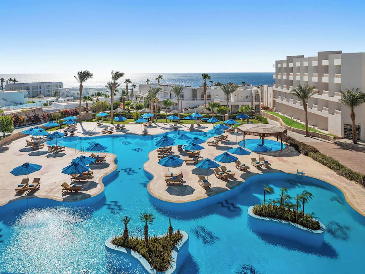 Aerial view of a resort with a large blue swimming pool, numerous blue umbrellas, lounge chairs, palm trees, and the ocean.