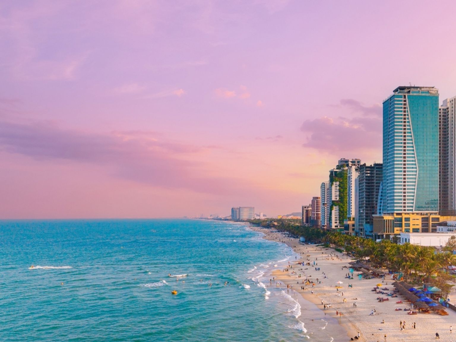 Luftfoto af en travl bystrand ved solnedgang, med høje bygninger langs kysten og mennesker på sandet og i det turkise vand. Da Nang - Vietnam