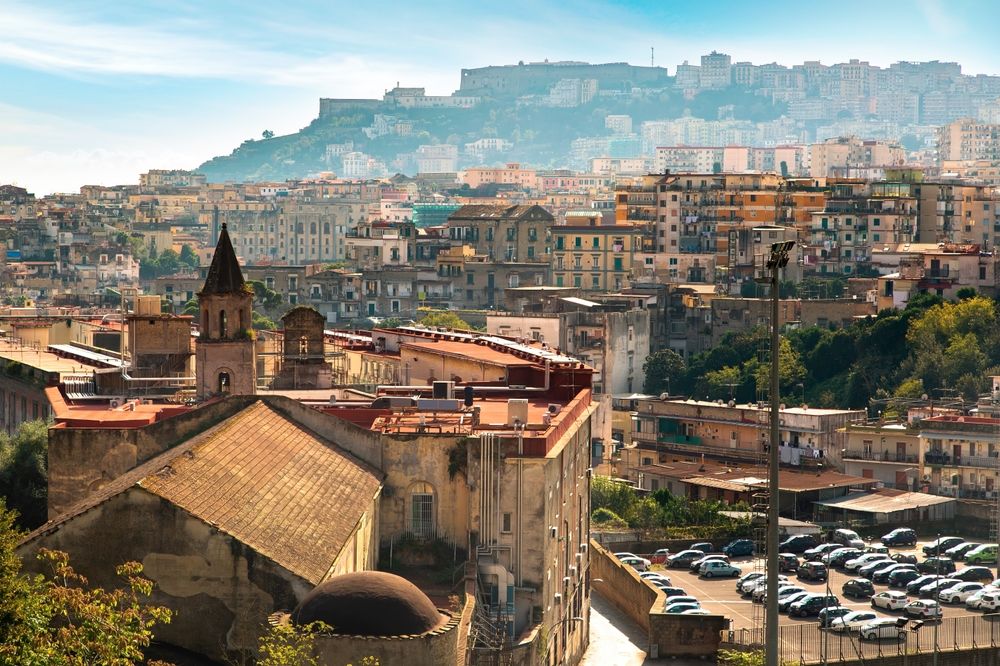 Paysage urbain d’une ville densément bâtie sur des collines, avec un clocher d’église au premier plan et une forteresse au sommet d’une colline lointaine. Naples, Italie