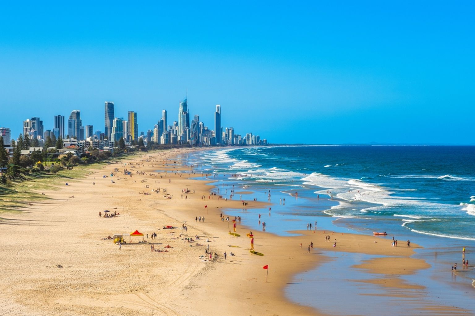 Overfyldt sandstrand med bølger og en fjern bysilhuet under en klar blå himmel. Brisbane