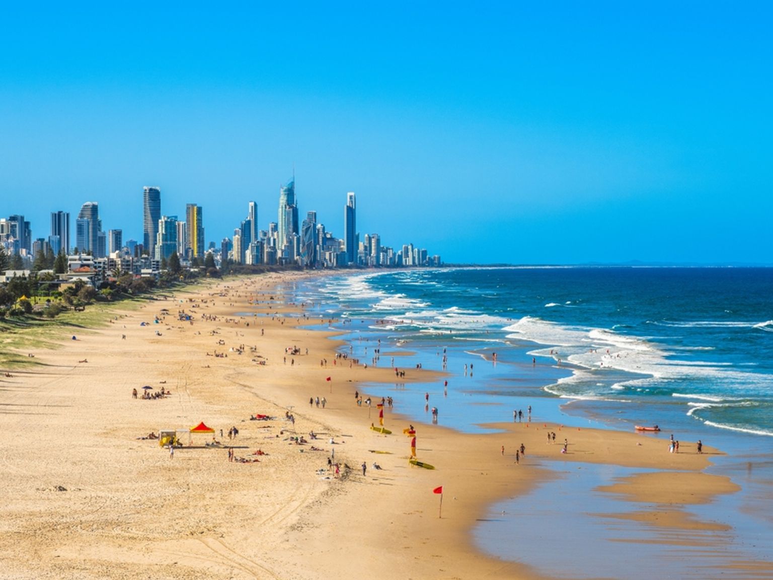 Trångt sandstrand med vågor och en avlägsen stadssilhuett under en klarblå himmel. Brisbane