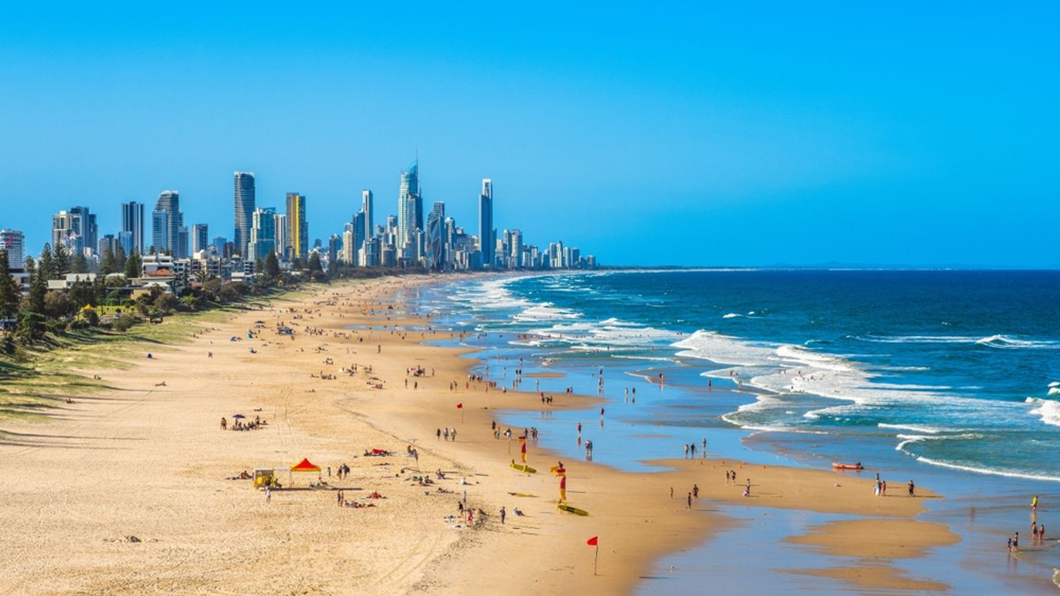 Overfyldt sandstrand med bølger og en fjern bysilhuet under en klar blå himmel. Brisbane