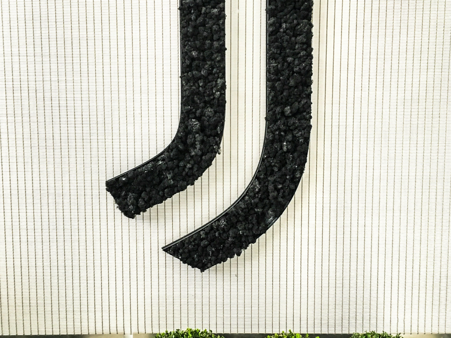 Das Juventus J-Logo, texturiert mit schwarzen Kieselsteinen, auf einer gestreiften weißen Wand über drei runden grünen Büschen.