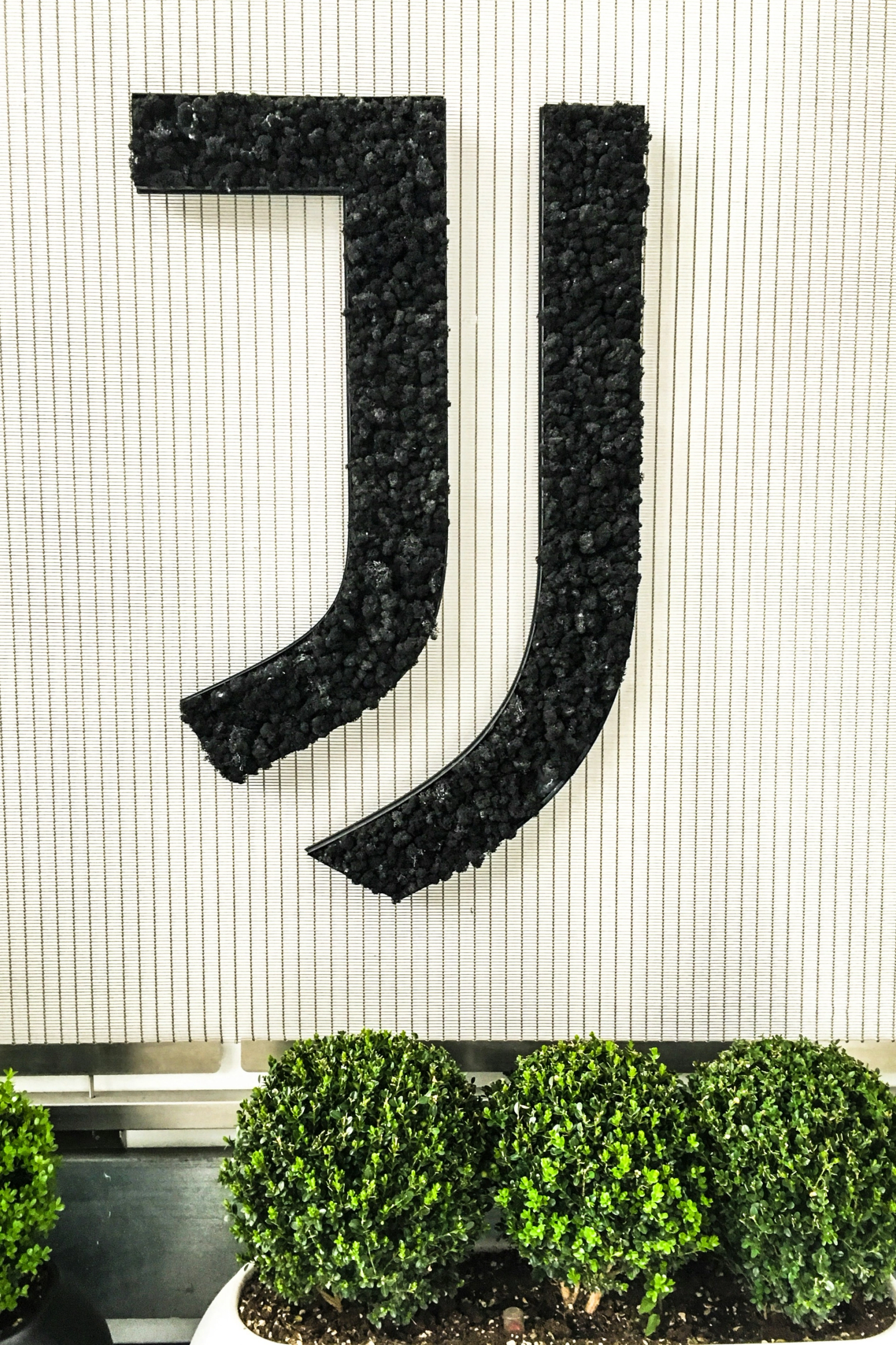 Das Juventus J-Logo, texturiert mit schwarzen Kieselsteinen, auf einer gestreiften weißen Wand über drei runden grünen Büschen.