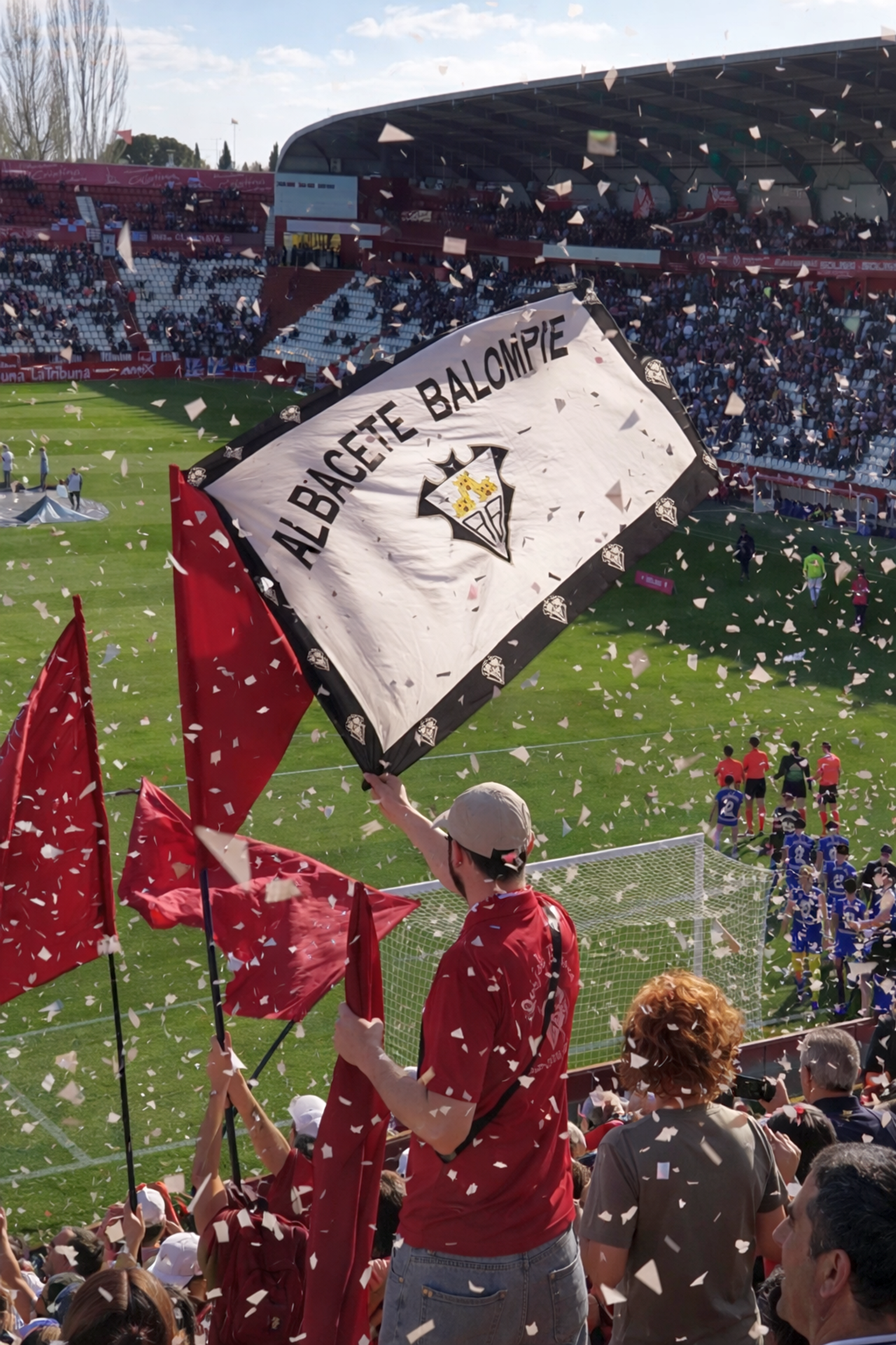 Torcedores agitam uma bandeira do "Albacete Balompié" e bandeiras vermelhas em um estádio de futebol repleto de confetes.