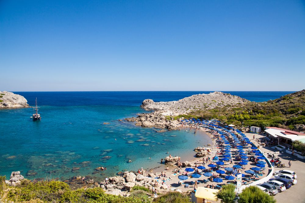 Scenic bay with a crowded beach under blue umbrellas, clear turquoise water, rocky shores, a boat, and a seaside restaurant.