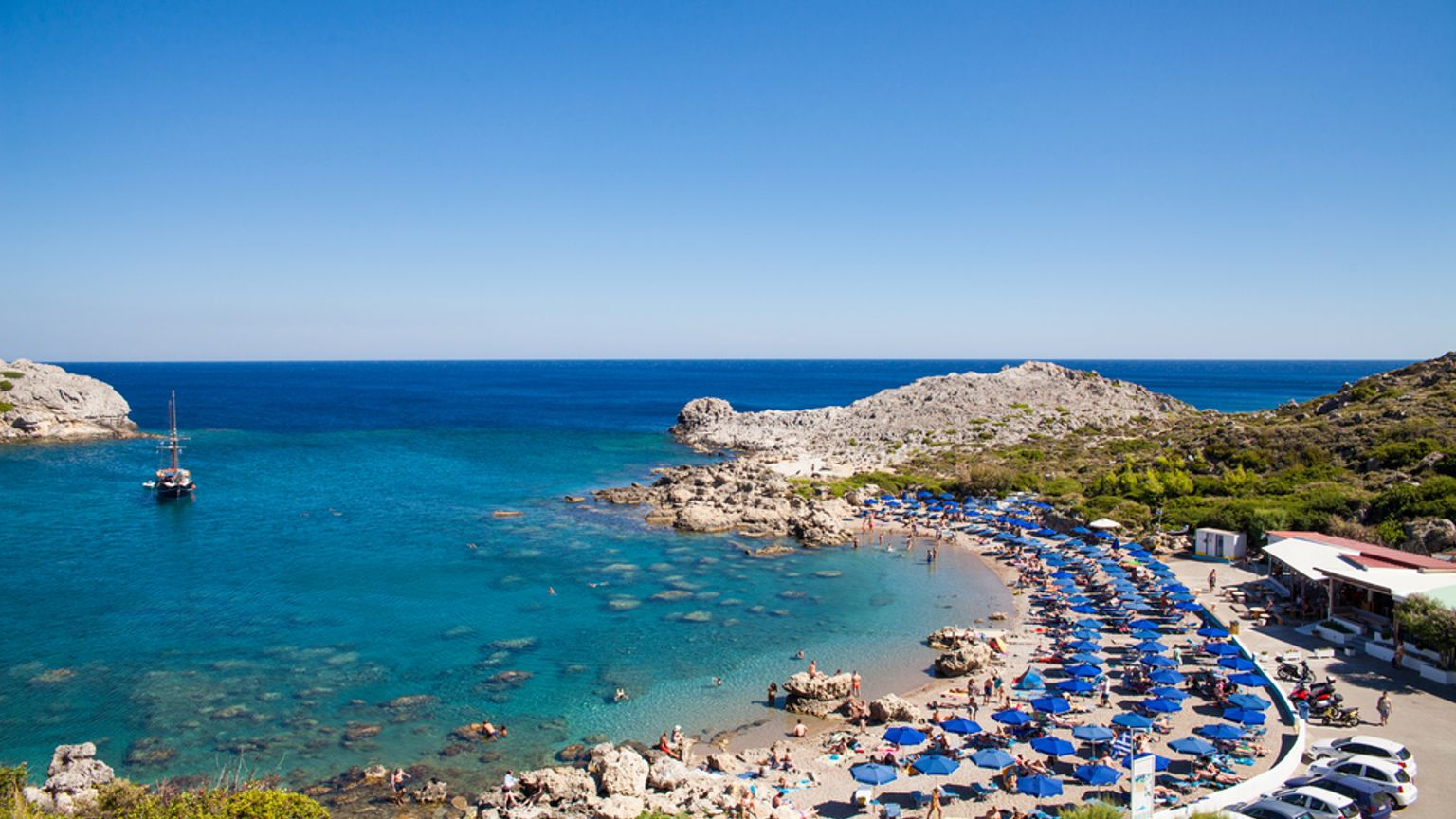 Baie pittoresque avec une plage bondée sous des parasols bleus, une eau turquoise et limpide, des rivages rocheux, un bateau et un restaurant en bord de mer.