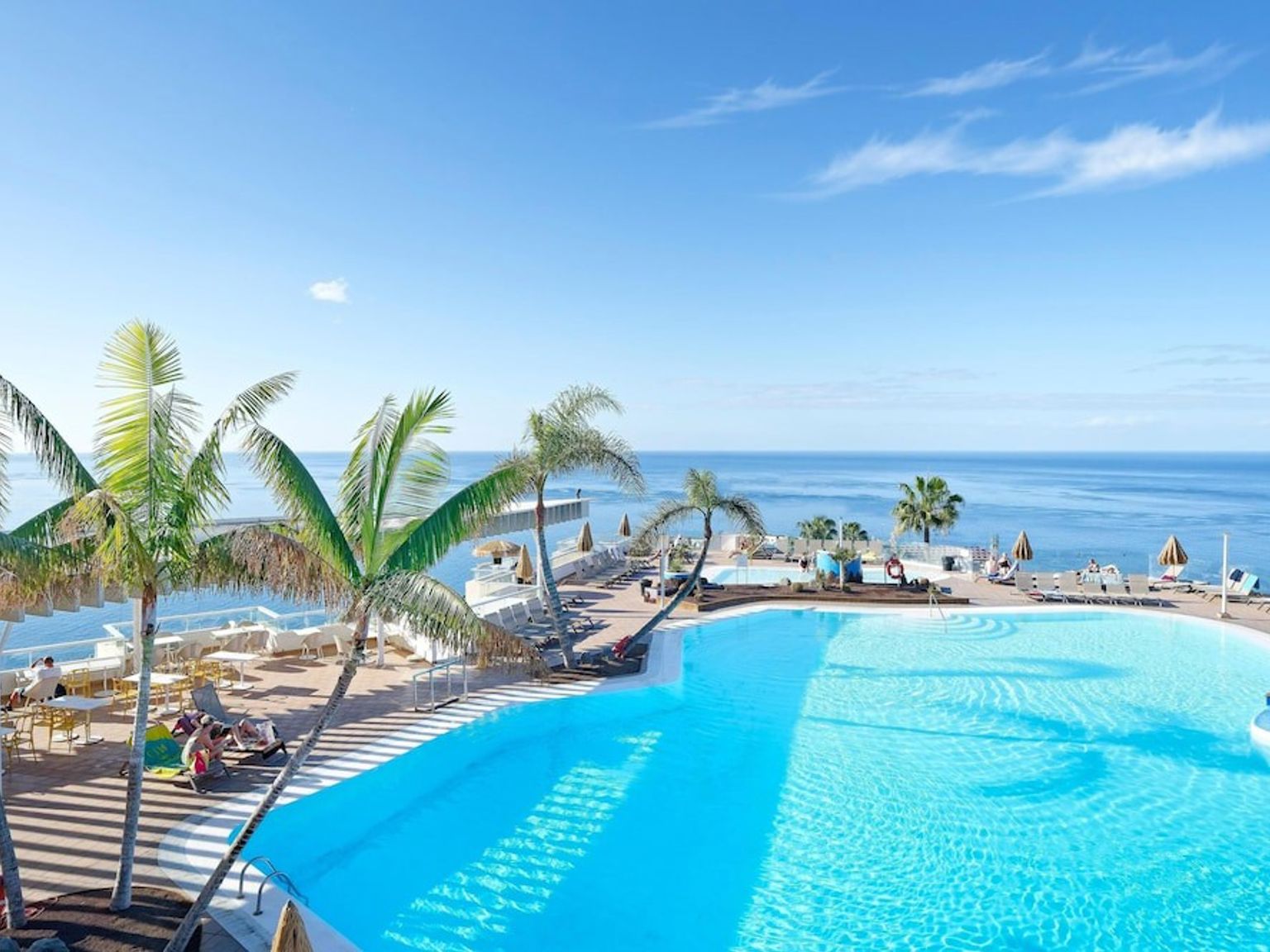 Resortpool med palmer, solbadare och havsutsikt under en klar himmel.