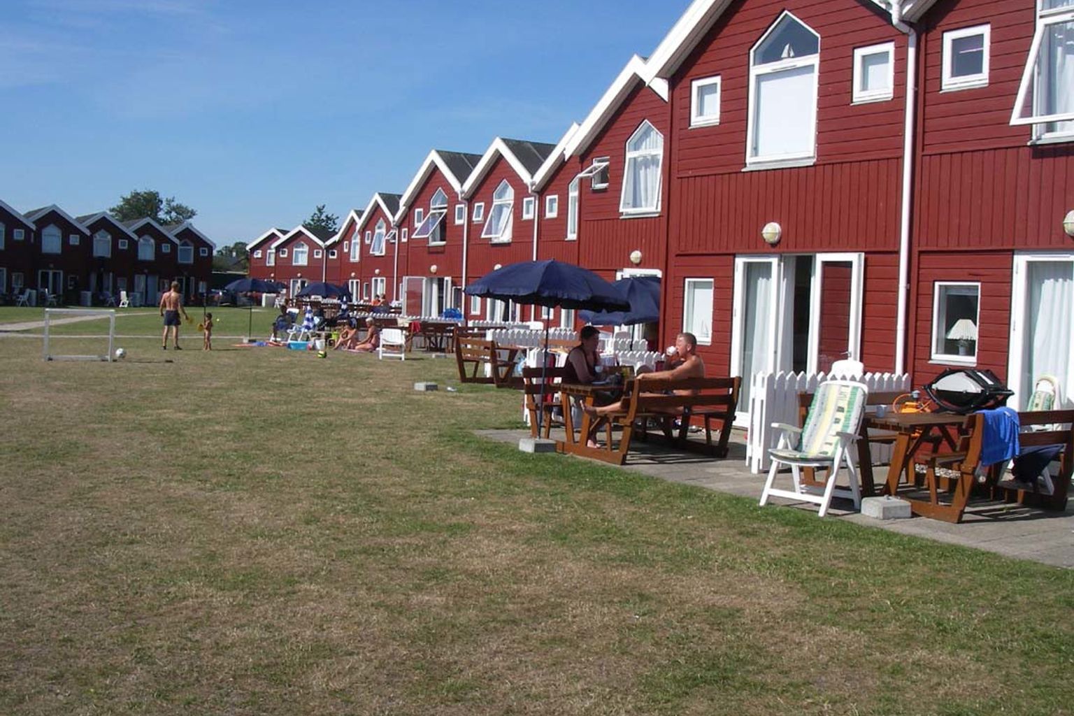 Røde huse ligger på række langs en solrig, tør græsmark, hvor folk slapper af på terrasser, og børn spiller fodbold.