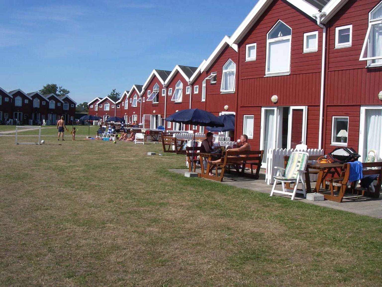Røde huse ligger på række langs en solrig, tør græsmark, hvor folk slapper af på terrasser, og børn spiller fodbold.