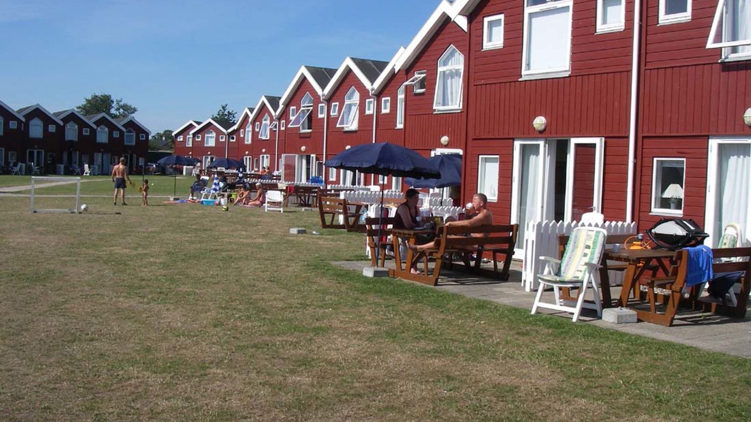 Røde huse ligger på række langs en solrig, tør græsmark, hvor folk slapper af på terrasser, og børn spiller fodbold.