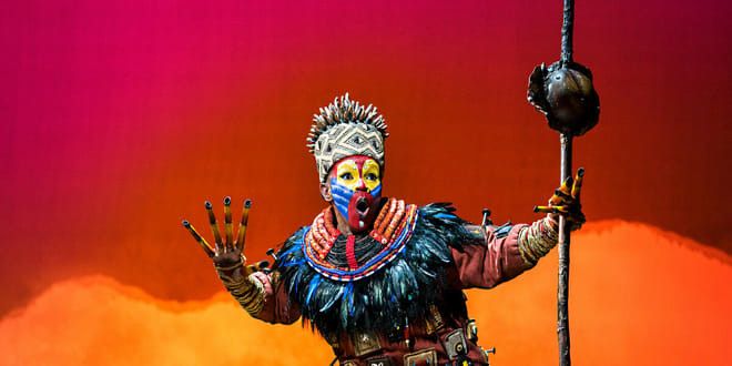 Artist i en utsmyckad Rafiki-kostym med färgglad ansiktsmålning och huvudbonad, som håller en stav mot en livlig röd och orange bakgrund.