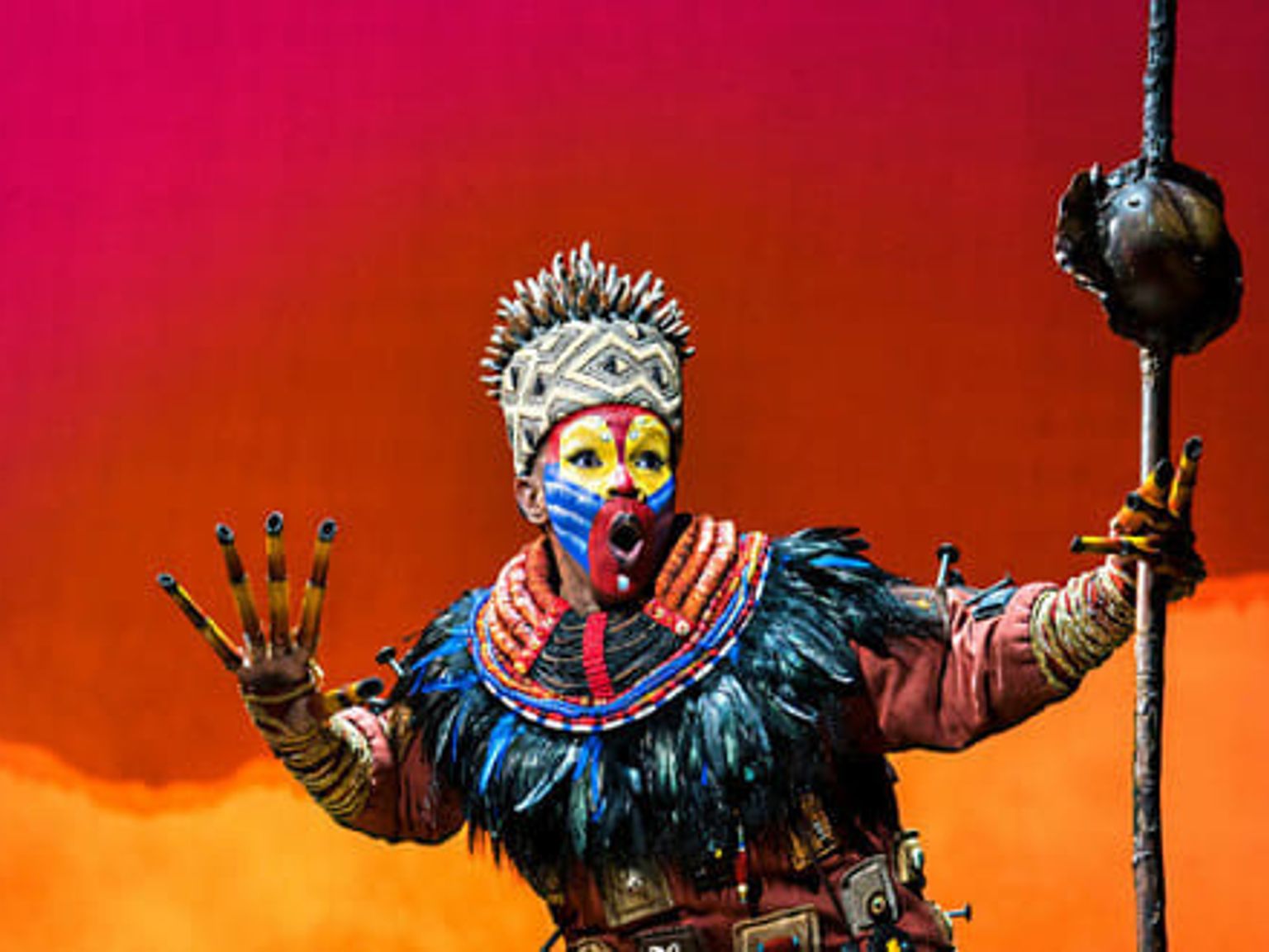 Artist i en utsmyckad Rafiki-kostym med färgglad ansiktsmålning och huvudbonad, som håller en stav mot en livlig röd och orange bakgrund.