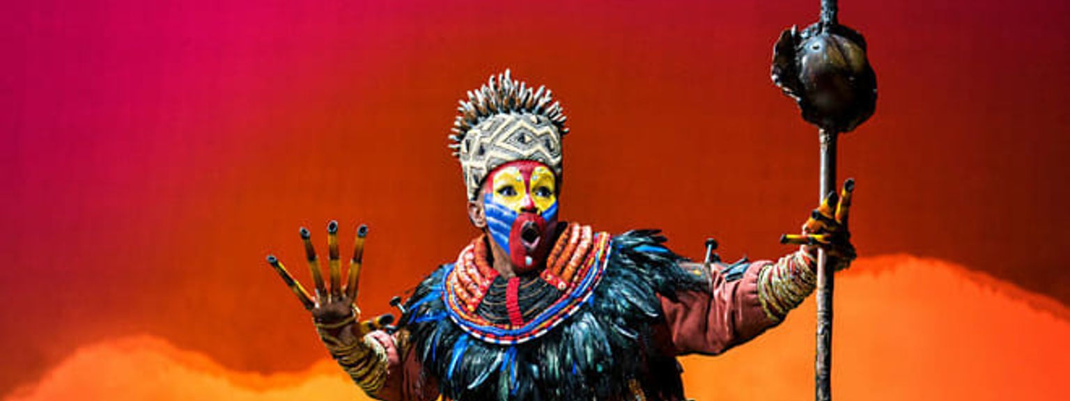 Artist i en utsmyckad Rafiki-kostym med färgglad ansiktsmålning och huvudbonad, som håller en stav mot en livlig röd och orange bakgrund.