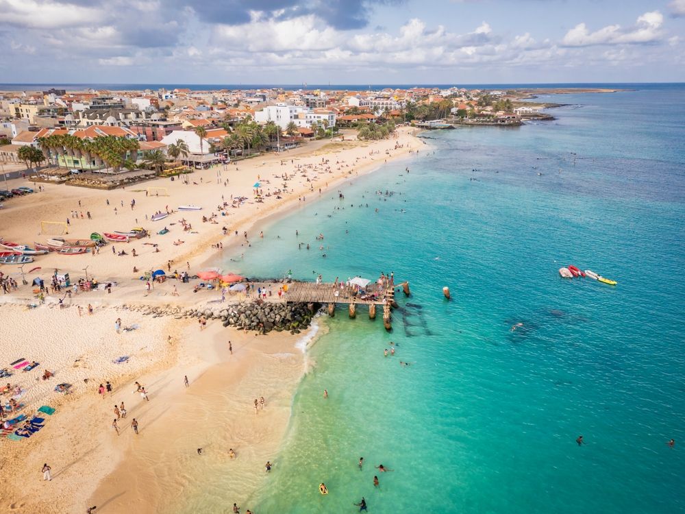 Luftfoto af en fyldt tropisk strand med turkis vand, en mole og en kystby.