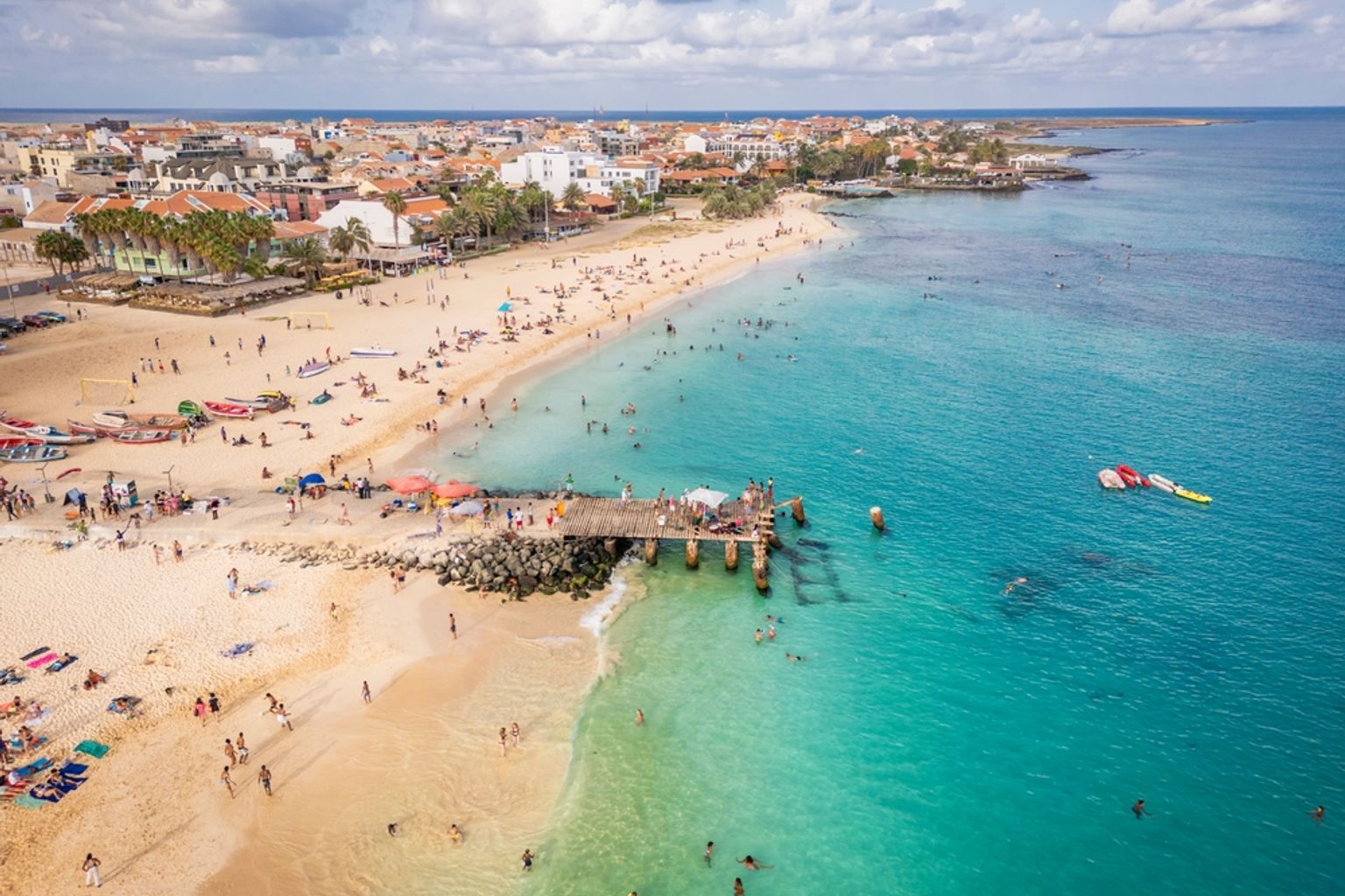 Luftaufnahme eines überfüllten tropischen Strandes mit türkisfarbenem Wasser, einem Pier und einer Küstenstadt.