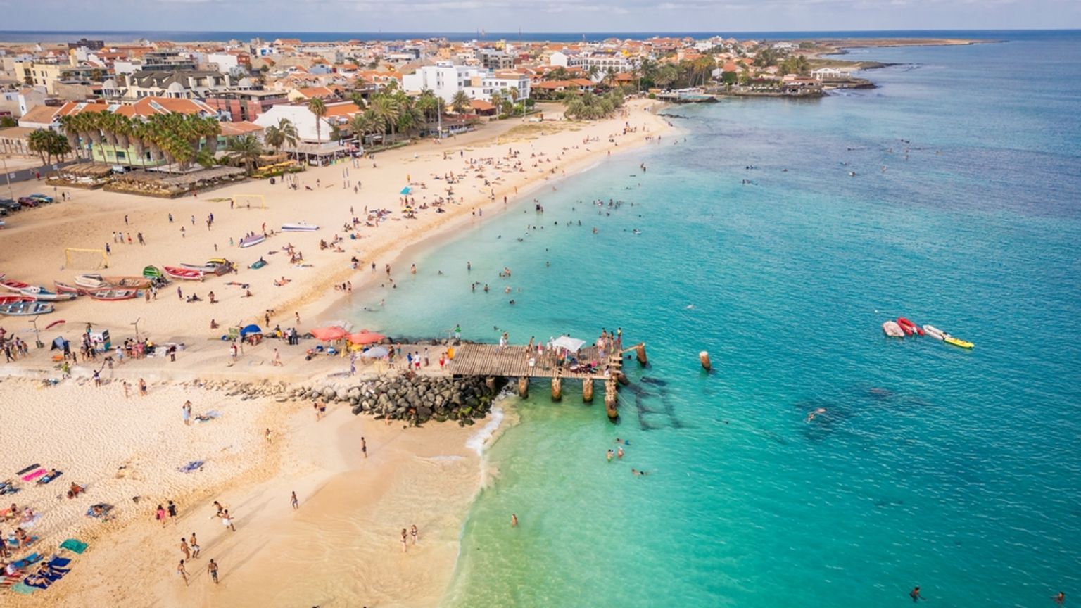 Luftaufnahme eines überfüllten tropischen Strandes mit türkisfarbenem Wasser, einem Pier und einer Küstenstadt.