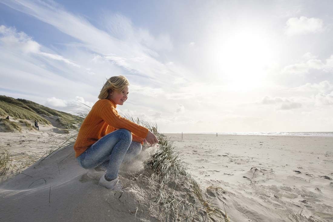 Ung jente i oransje genser som sitter på huk på en sanddyne på stranden. Landal Grønhøj Strand
