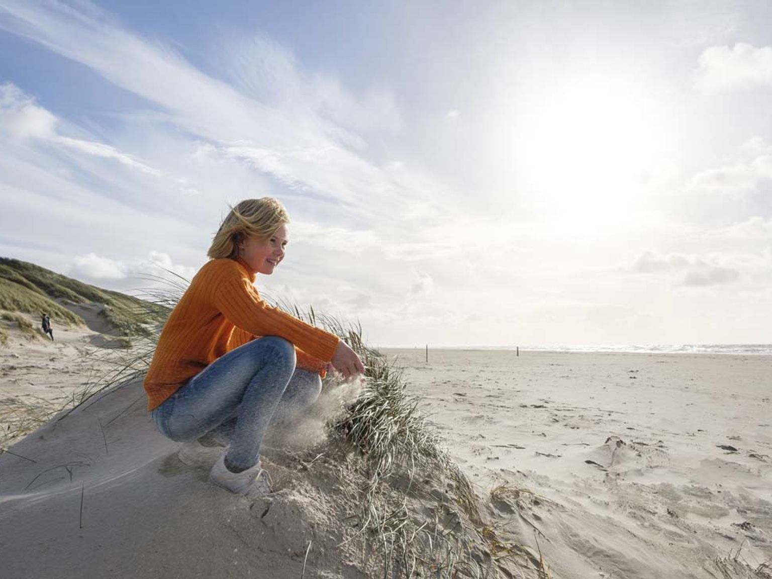 Ung jente i oransje genser som sitter på huk på en sanddyne på stranden. Landal Grønhøj Strand