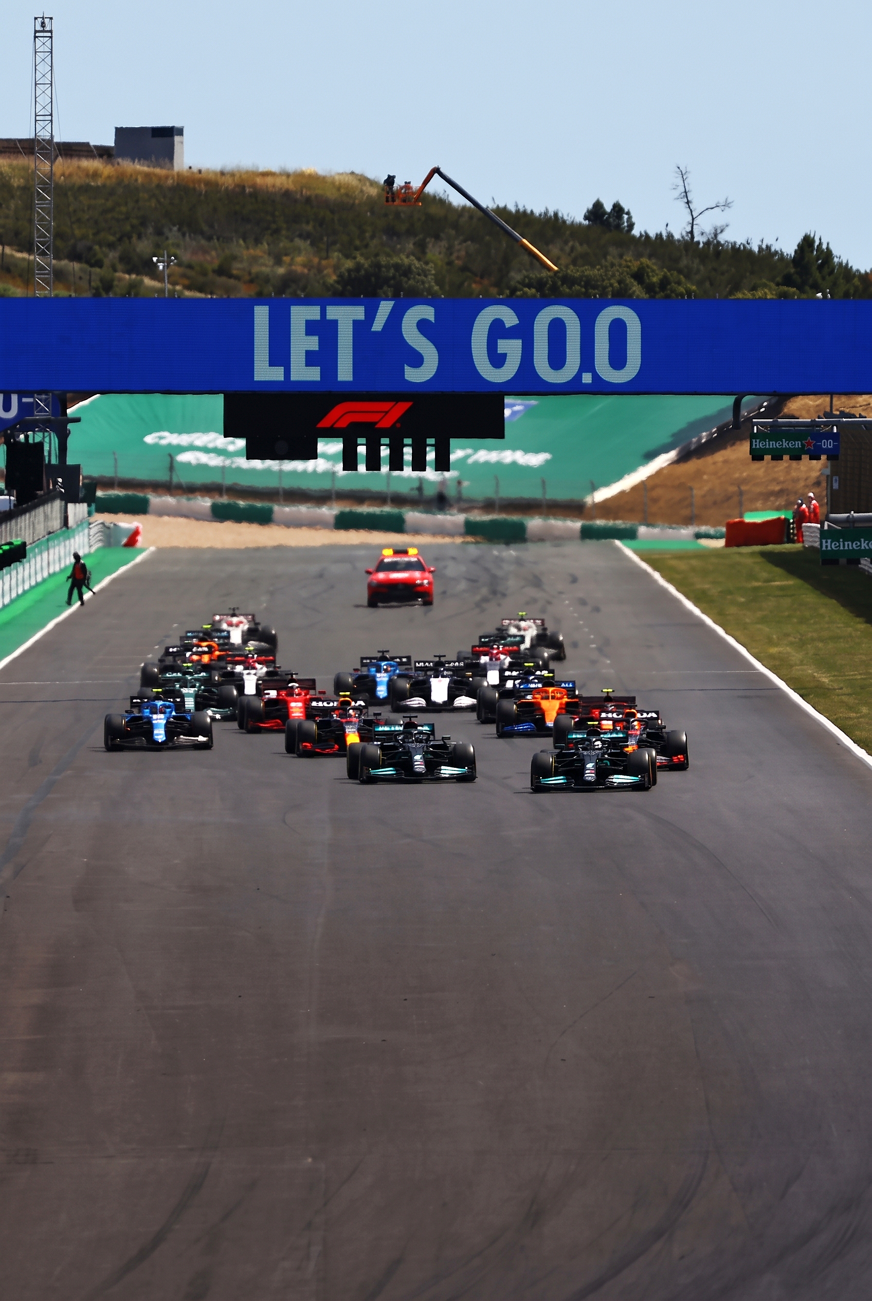 Een veld Formule 1-auto's volgt een safetycar op een circuit, met daarboven een spandoek met de tekst 'LET'S GO'.