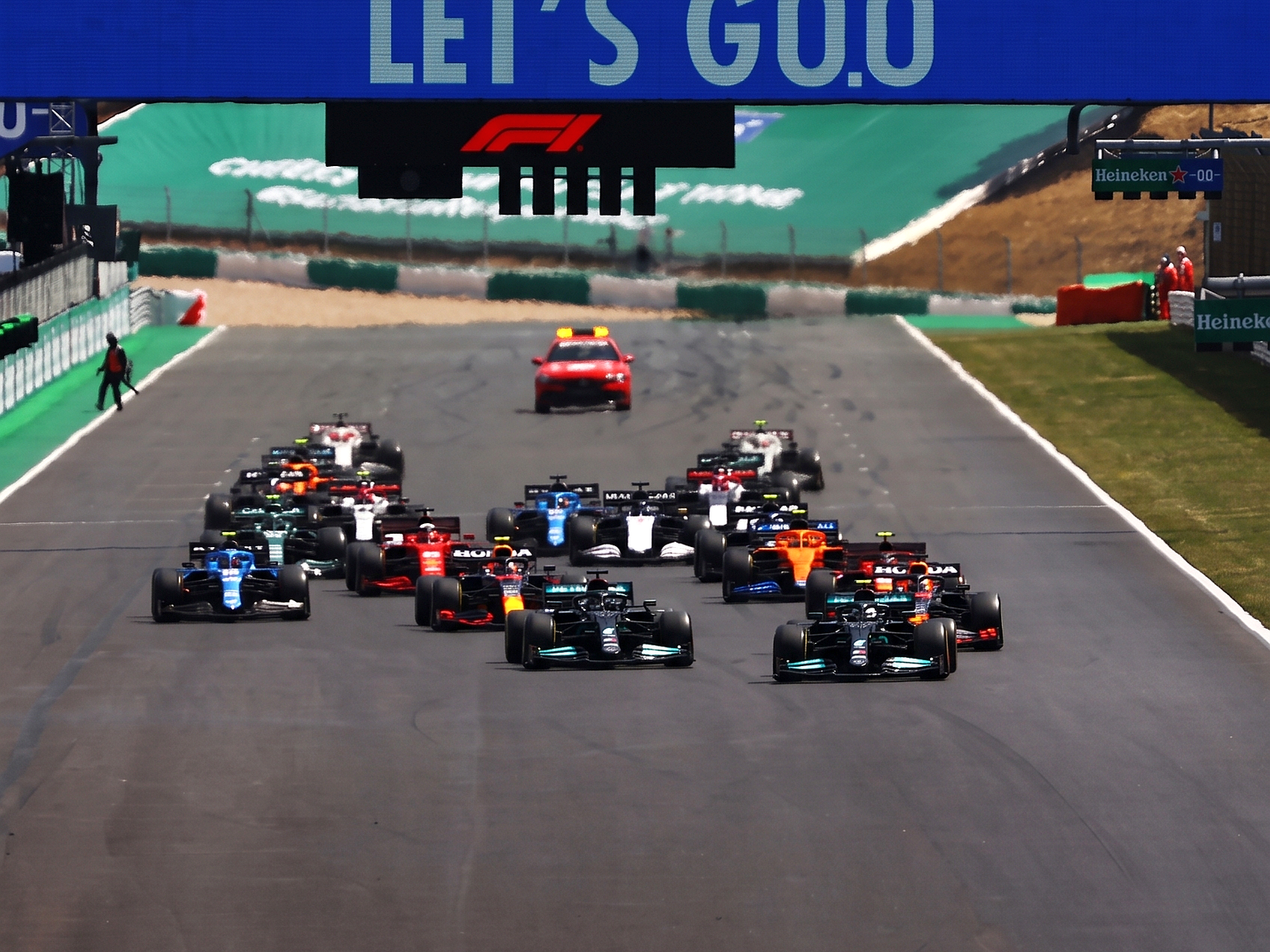 Een veld Formule 1-auto's volgt een safetycar op een circuit, met daarboven een spandoek met de tekst 'LET'S GO'.