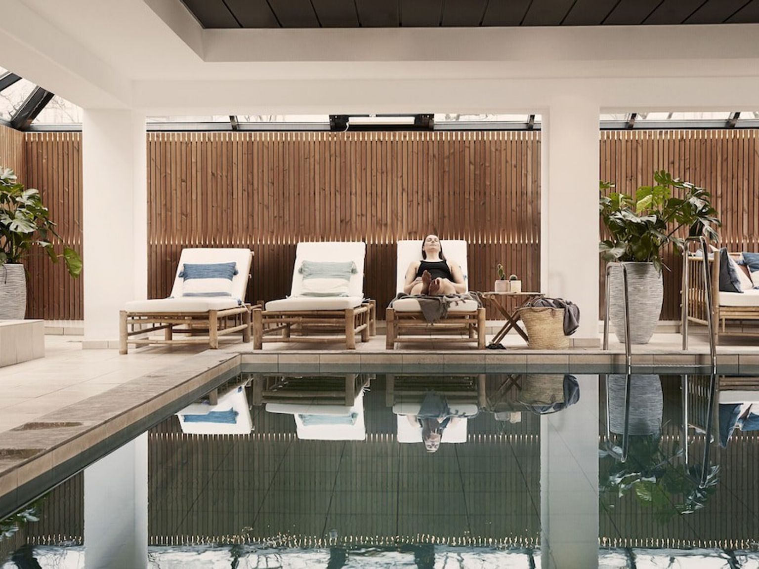En kvinna kopplar av på en solstol vid en inomhuspool, omgiven av moderna träväggar och krukväxter.