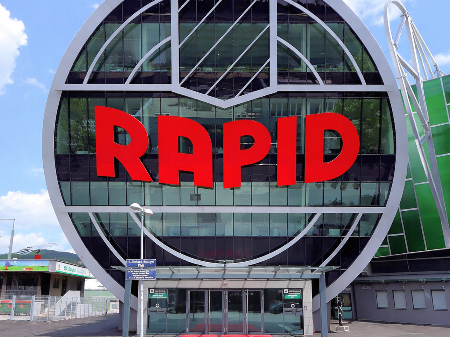 Moderne Gebäude-Eingang mit einer runden Glasfassade, die "RAPID" in Rot anzeigt, gekrönt von drei weißen Sternen.