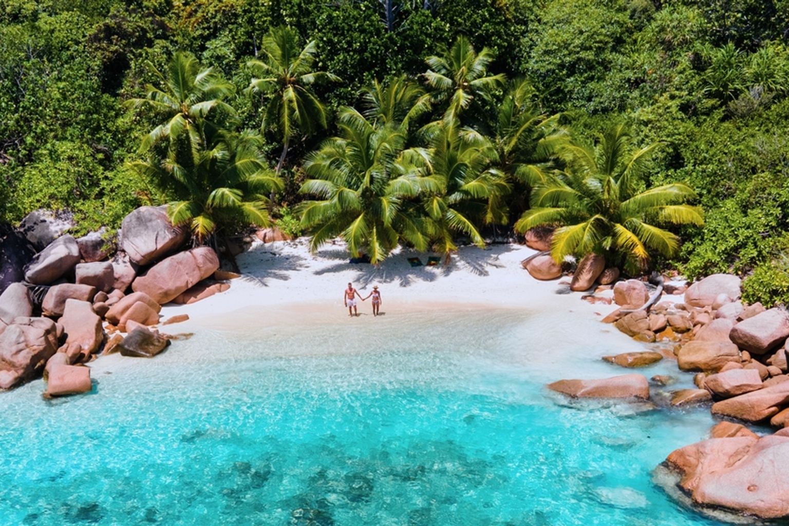 Flygfoto av ett par som håller varandra i handen på en vit sandstrand med turkost vatten, omgiven av stora klippblock och frodiga gröna palmer. Praslin Island, Seychellerna.