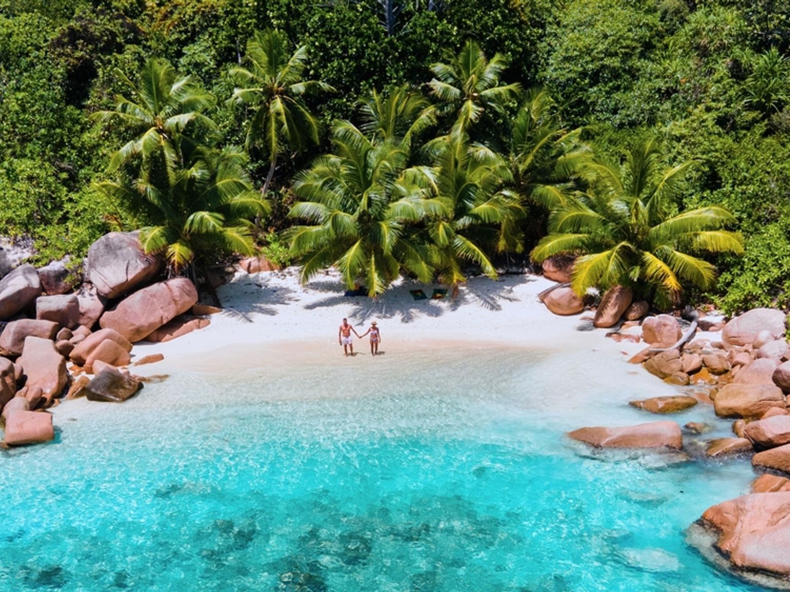 Flygfoto av ett par som håller varandra i handen på en vit sandstrand med turkost vatten, omgiven av stora klippblock och frodiga gröna palmer. Praslin Island, Seychellerna.