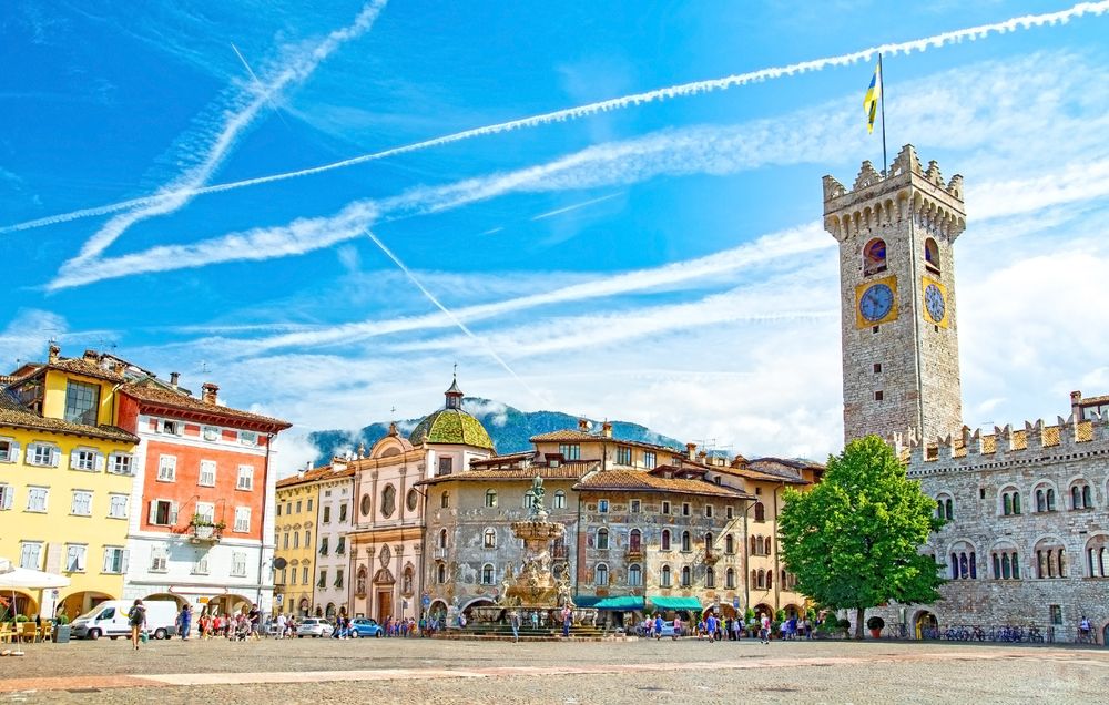En livlig europeisk torgplass med et høyt klokketårn i stein, fargerike historiske bygninger og mennesker under en klar blå himmel. Trento, Italia