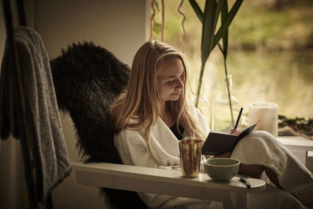 En kvinna i en vit morgonrock sitter i en fåtölj med päls vid ett fönster och skriver i en anteckningsbok.