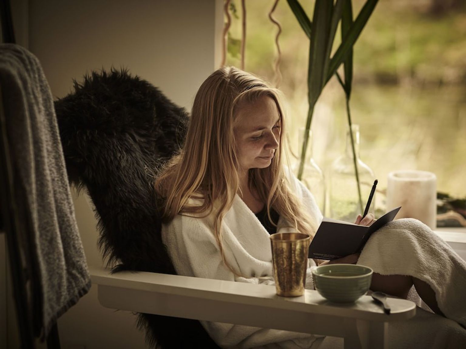 En kvinna i en vit morgonrock sitter i en fåtölj med päls vid ett fönster och skriver i en anteckningsbok.