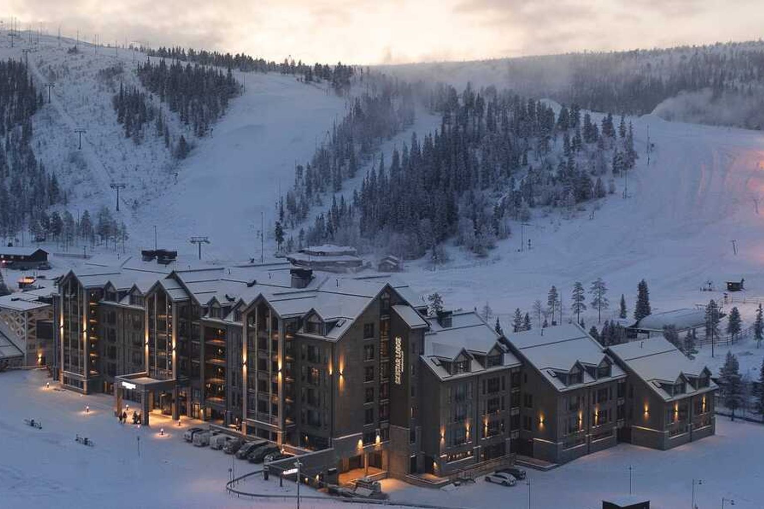 Et stort hotel med oplyste vinduer beliggende i en snedækket bjergskisportssted ved skumring.